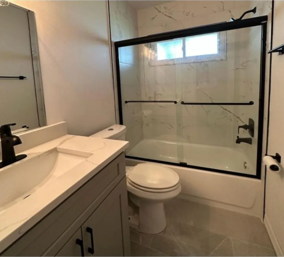 After: Bathroom after the remodel: teal tile tub and toilet replaced with white tile, new vanity and black framed glass doors