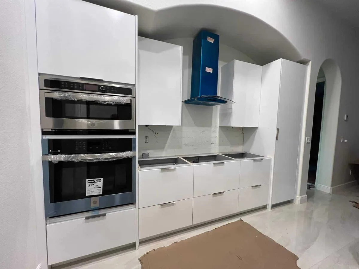 White slab cabinets with wall ovens and a blue chimney hood during installation